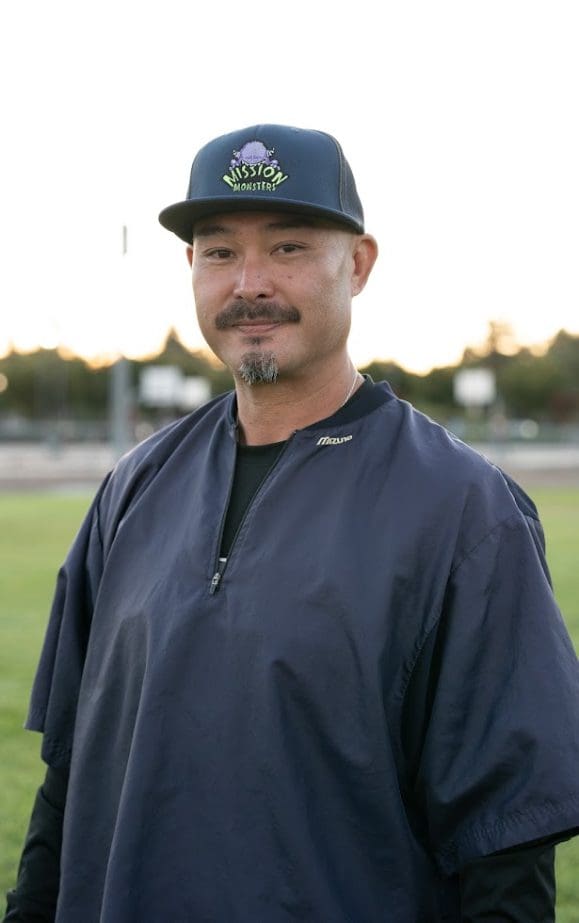 Man smiling outside wearing a navy blue jacket and a baseball cap.